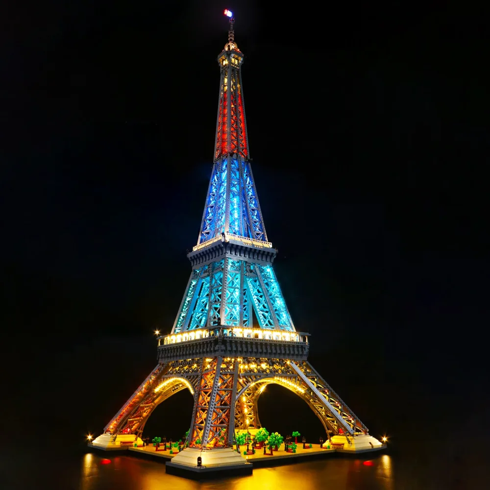The LEGO Architecture Eiffel Tower lit in the dark, red white and blue through the upper shaft, warm gold along the arches and base, and a small beacon at the summit