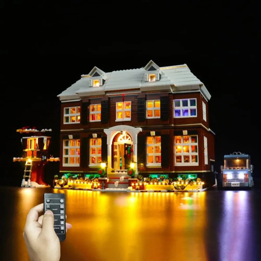 The LEGO Ideas Home Alone house lit at night with warm window light and a run of small lights along the front path, a hand holding the kit's small remote control in the foreground