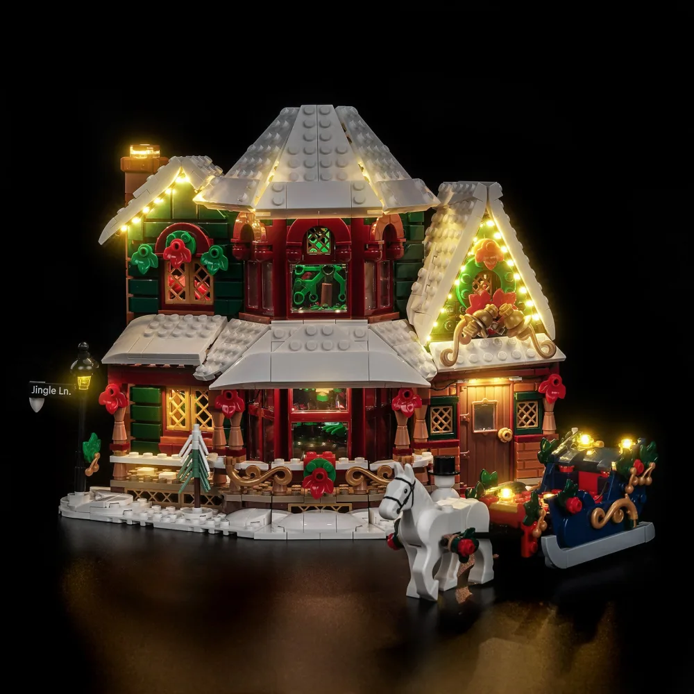A LEGO Christmas house lit at night, warm light in the windows, a string of lights along the rooflines, a lit street lamp marked Jingle Lane and a lit sleigh beside it