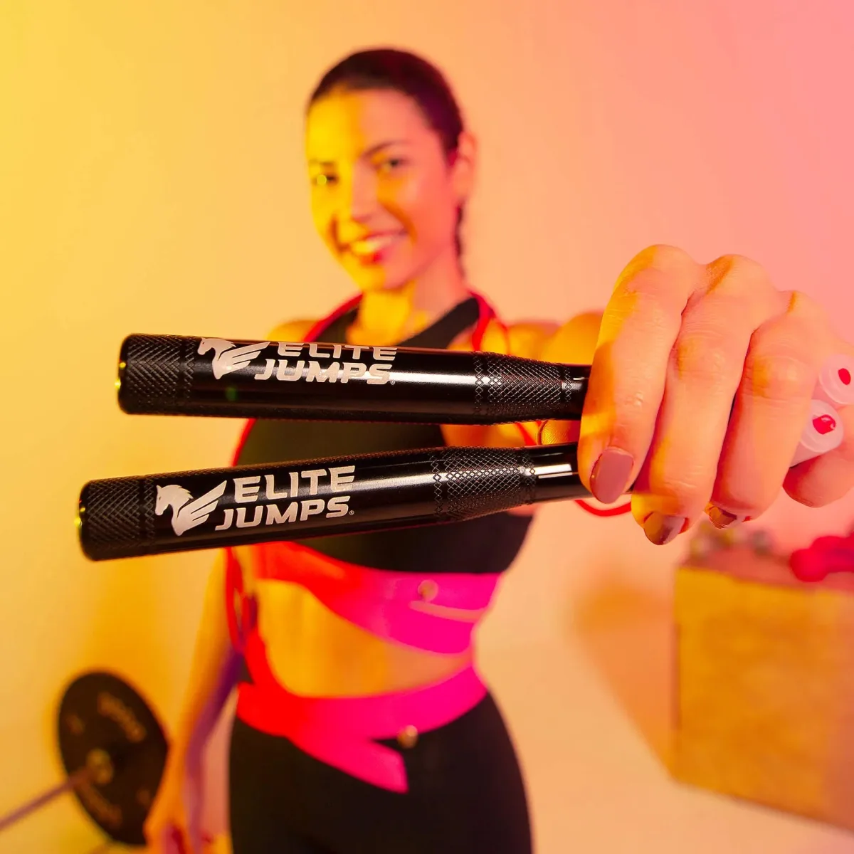 Best Jump Ropes for Double Unders: Speed Ropes, Cables and Sizing (2026) 1 Close view of two black knurled Elite Jumps speed rope handles held out toward the camera, with a woman in gym clothing smiling out of focus behind them