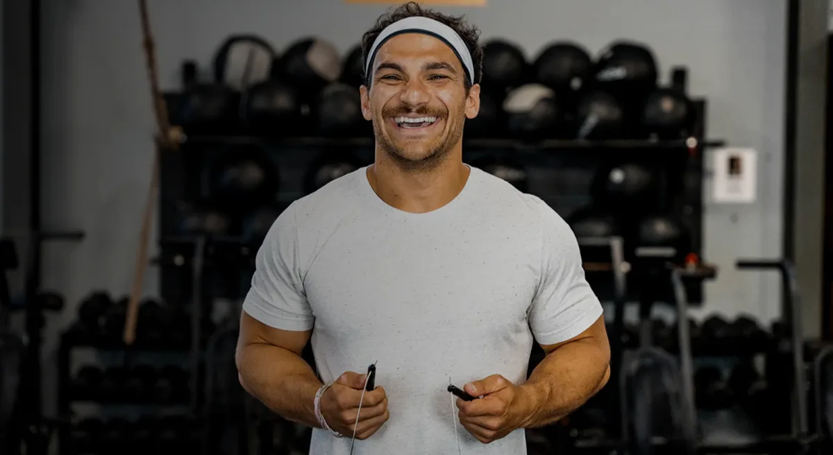 Best Jump Ropes for Double Unders: Speed Ropes, Cables and Sizing (2026) 2 A man in a headband and grey t-shirt standing in a gym, holding a speed rope handle in each hand with racks of medicine balls and dumbbells behind him