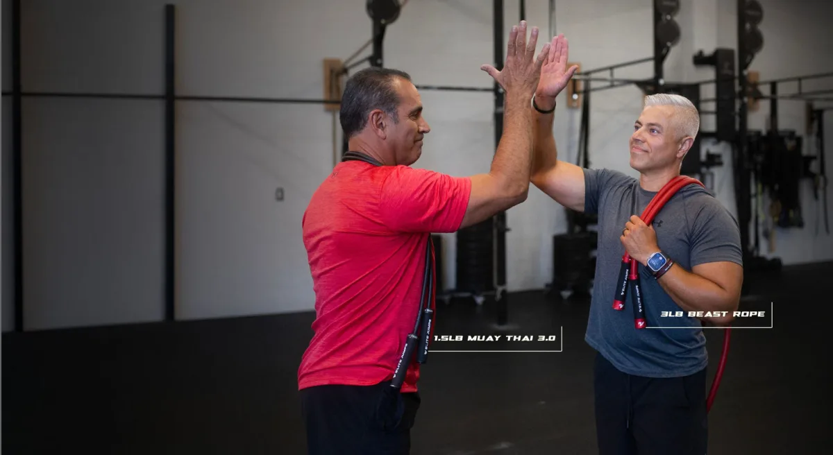 Best Jump Ropes for Double Unders: Speed Ropes, Cables and Sizing (2026) 3 Two men high-fiving in a gym, one with a 1.5lb Muay Thai 3.0 rope over his shoulders and the other with a red 3lb Beast rope, each labelled on the image