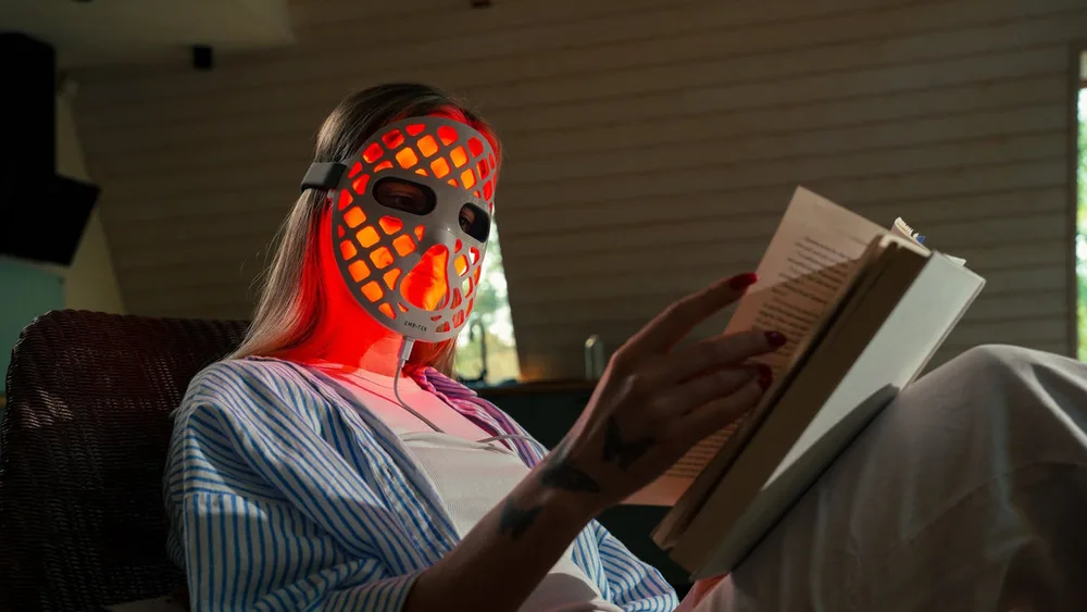 A woman reads a book while wearing the EMR-TEK ETERNO LED face mask, a rigid lattice mask glowing red with open eye holes