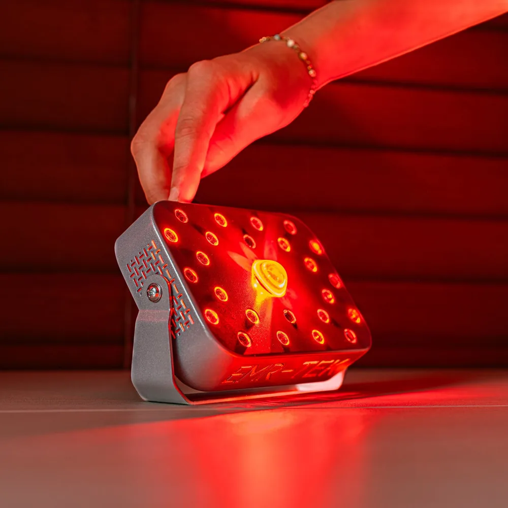 Close-up of the EMR-TEK Firefly, a compact grey device with rows of lit red emitters and the brand name on its front edge, being adjusted by hand