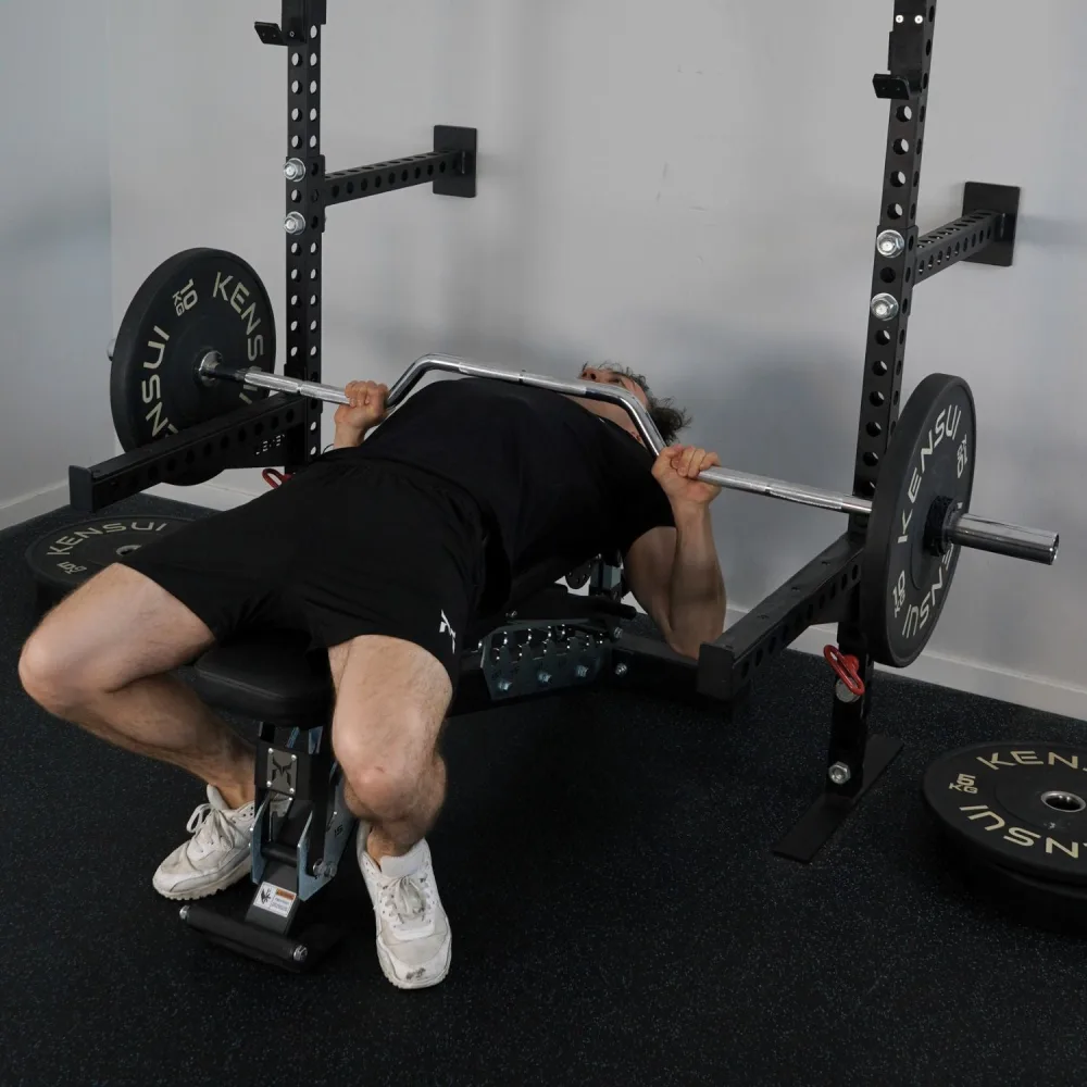Best Specialty Barbells for Home Gyms: Trap, Swiss, Cambered and Buffalo Bars Compared 4 A man bench pressing in a rack with the Kobe Cambered Bar, whose handles drop below the line of the shaft to let the hands travel lower than the chest