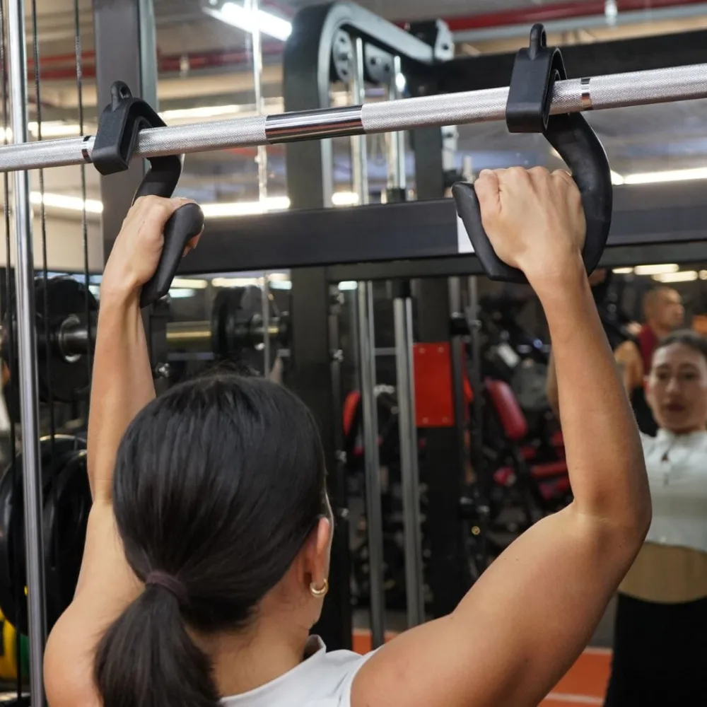 A woman doing pull-ups in a gym, gripping two black Swissies handles hooked over a straight bar rather than the bar itself