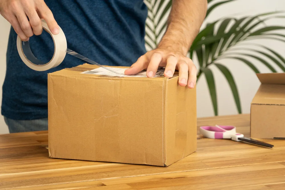 Một người mặc áo sơ mi xanh đang dùng băng keo dán kín một thùng carton màu nâu trên một chiếc bàn gỗ, bên cạnh là chiếc kéo và một thùng rỗng khác.