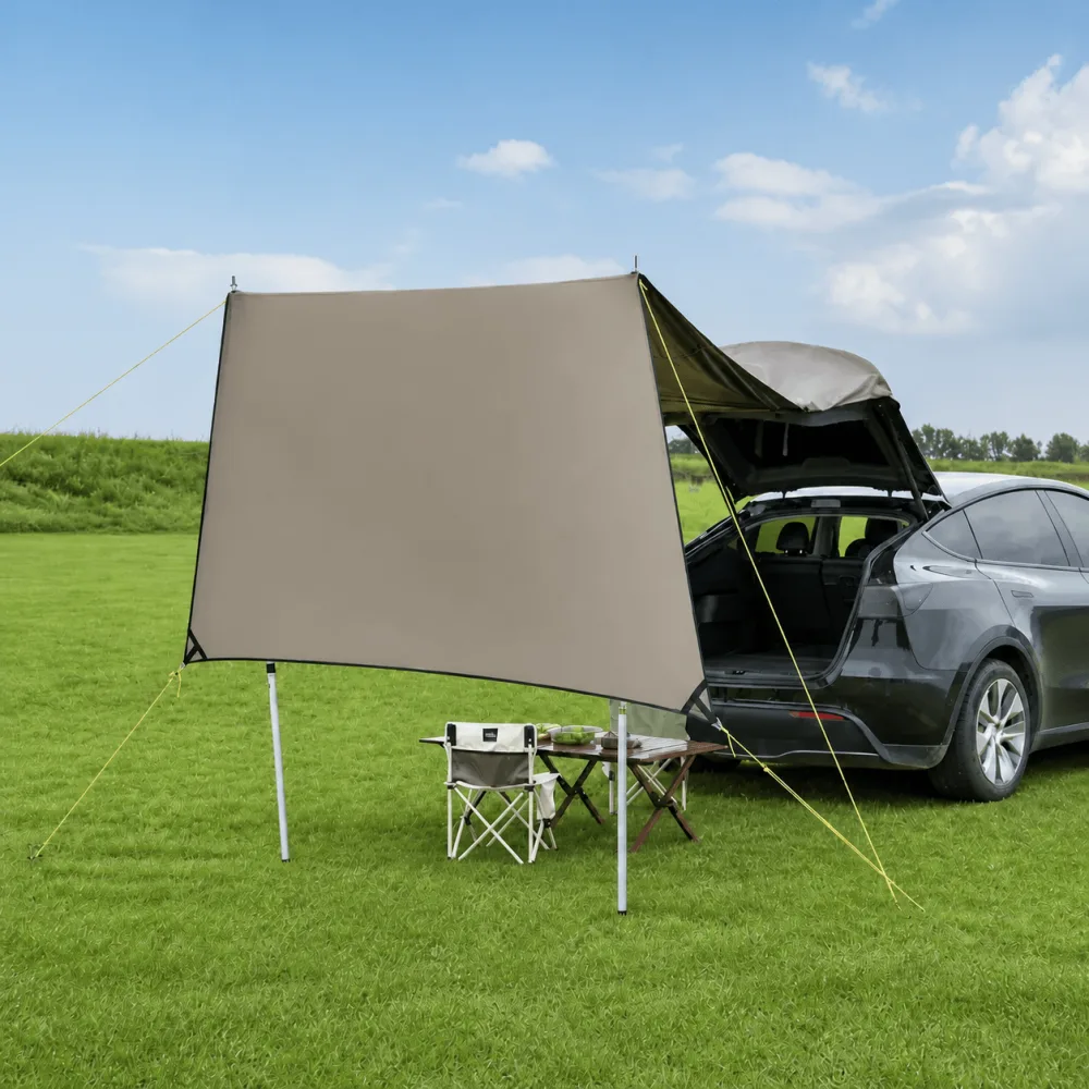 A fabric awning pitched from the open tailgate of a grey Tesla Model Y on grass, with a folding table and chair underneath